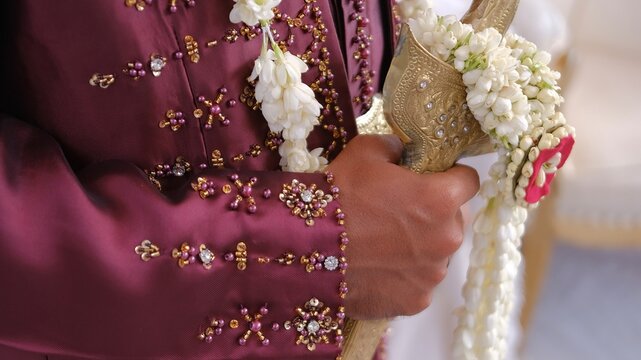 Detailed shot of a groom in elegant maroon wedding attire with intricate gold and purple beadwork embroidery, holding a golden heritage dagger.
