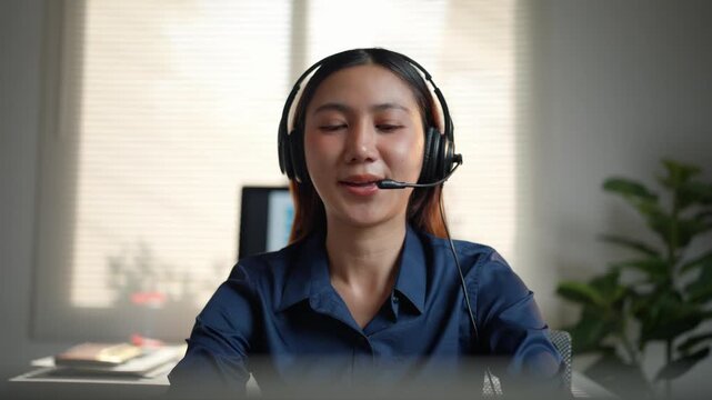 Smiling young Asian woman wearing a headset, working in a call center, speaking with a client. Customer service representative offering support