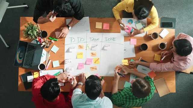 Top down aerial view of diverse business team making scrum task board at meeting room. Group of smart people writing at paper and sticky notes for making kanban board to manage work flow. Convocation.