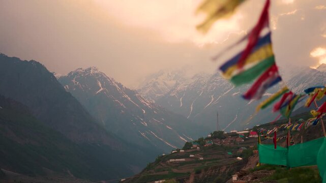4K Landscape shot of orange clouds surrounding the snowy Himalayan mountain peak during a stormy weather with defocused Buddhist prayer flags waving in the front as seen from Lahaul in Himachal, India
