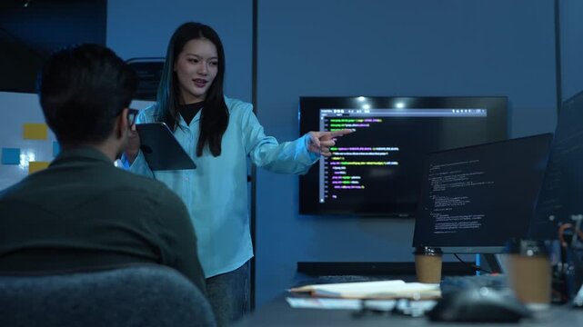Asian female manager talking to male programmer writing code in dark blue office, Tech team discussing software development, cybersecurity and AI technology, Late night working business team concepts