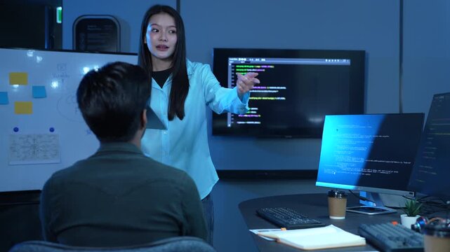 Asian female manager talking to male programmer writing code in dark blue office, Tech team discussing software development, cybersecurity and AI technology, Late night working business team concepts