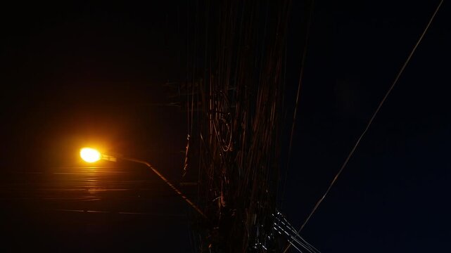 Dramatic low angle night view of tangled electric power cable wires on utility pole with glowing orange street lamp against dark blue sky