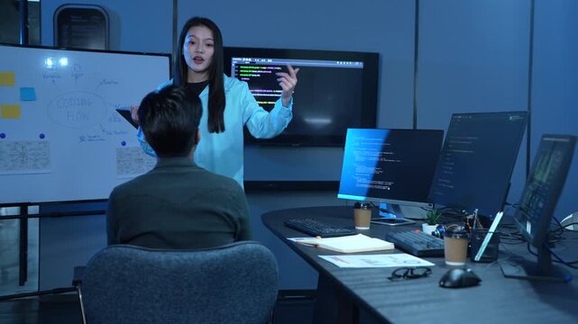 Asian female manager talking to male programmer writing code in dark blue office, Tech team discussing software development, cybersecurity and AI technology, Late night working business team concepts