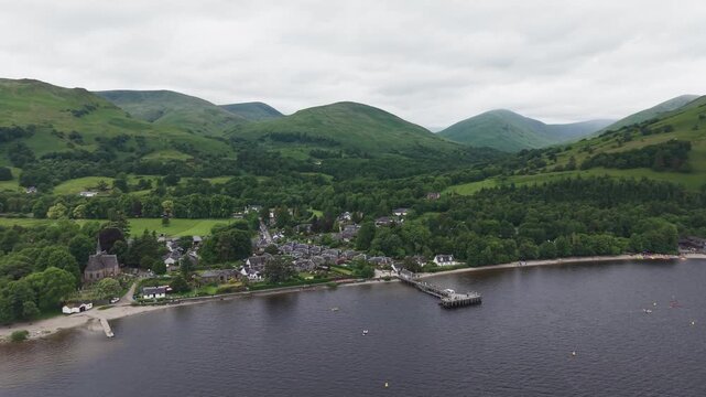 Aerial forward view of Luss village at Loch Lomond, Scottish Highlands. Drone flying over the lake towards the scenic shoreline with a historic pier, church, and green mountain slopes.