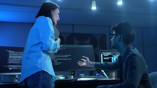 Asian female manager talking to male programmer writing code in dark blue office, Tech team discussing software development, cybersecurity and AI technology, Late night working business team concepts