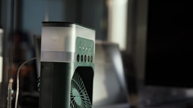 Close up of a portable desktop air cooler fan with a water tank sitting on a dark workspace desk next to a laptop and monitor in moody indoor lighting.