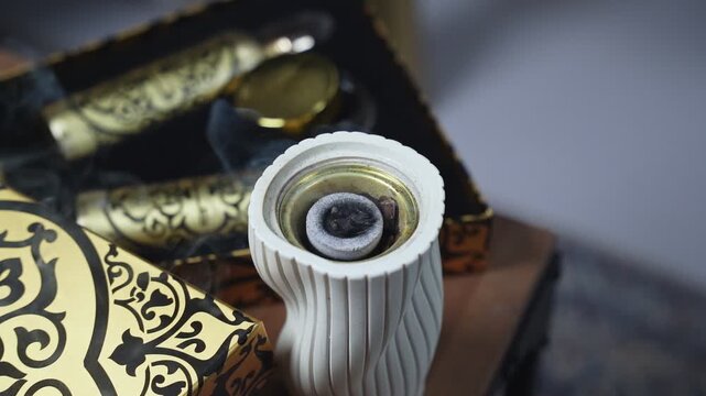 High angle close up of glowing charcoal burning bakhoor inside a white ceramic burner with smoke rising next to luxury gold patterned boxes.