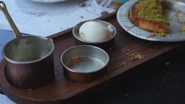 Close up of traditional middle eastern kunafa dessert topped with pistachio, served next to ice cream and a small syrup pot on a wooden tray at a restaurant.
