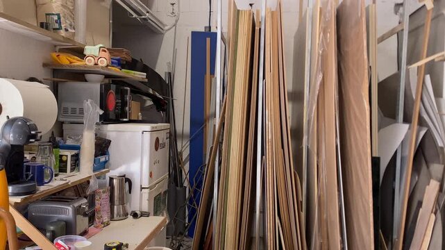 Messy storage room with stacked wood panels, shelves storing coffee machine, toaster, paper towels, and kitchen supplies