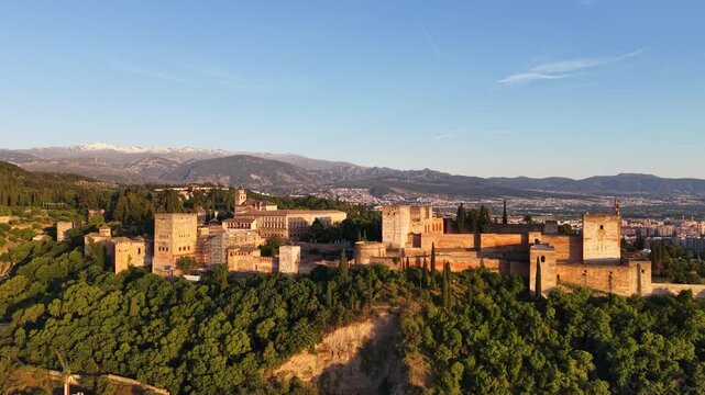 Drone aerial view of the Alhambra in Granada, Spain during golden hour with warm sunlight on the fortress and snow-capped Sierra Nevada mountains