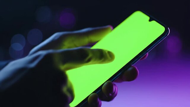 Close-up of Hands Using Smartphone with Green Screen Chroma Key in Neon Night Lighting ideal for app demonstrations and tech presentations.