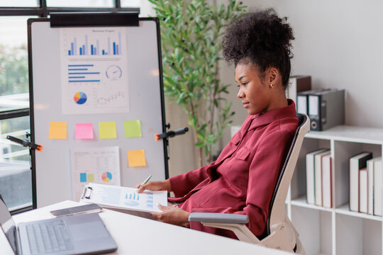 Young black businesswoman analyzing financial market data and charts on a clipboard, discussing business strategy, and presenting project statistics during an office meeting