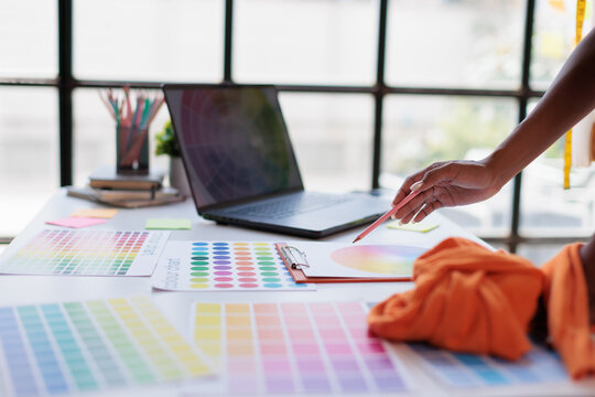 Designer's hand holding a pencil, selecting colors from a color wheel chart and swatches on a desk with a laptop, working on a creative design project