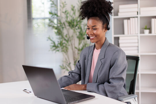 Black woman wearing headset and smiling while working at her laptop in a modern office. Providing excellent customer support in a call center. Managing virtual meetings