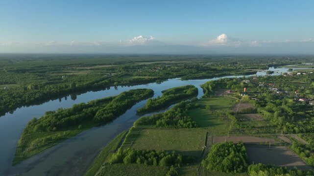 High angle drone shot of Anaklia village surrounded by lush green forests, agricultural fields, and a winding river under bright daylight.