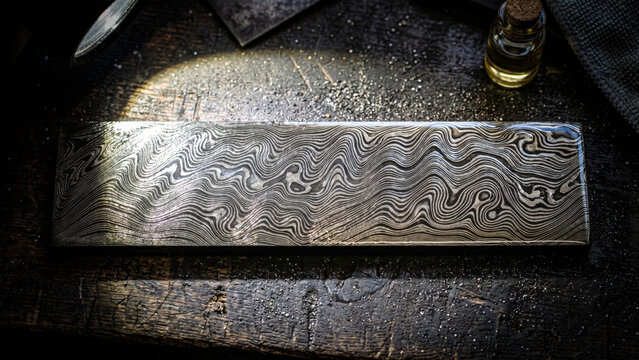 Macro top view of a layered Damascus steel sample bar revealing wavy acid etched watermark patterns and a high gloss protective oil sheen shimmering under a directional spotlight