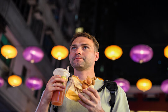 Handsome 30 years old Russian man tourist eating traditional bubble waffle and holding iced lemon tea at Temple Street Night Market in Hong Kong