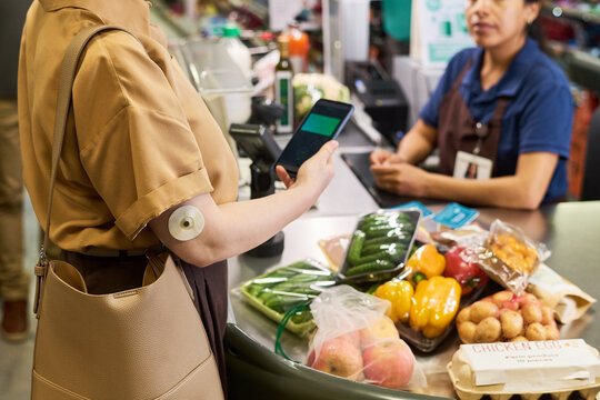 Adult woman with diabetes paying by smartphone at supermarket checkout, managing grocery shopping and glucose monitoring. Ideal for nutrition, chronic care, retail health