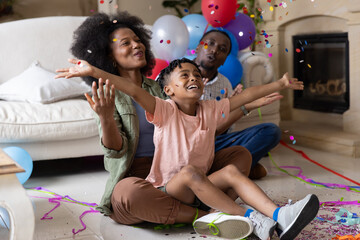 Obraz premium African American family sitting on floor in living room with balloons, confetti and smiling child