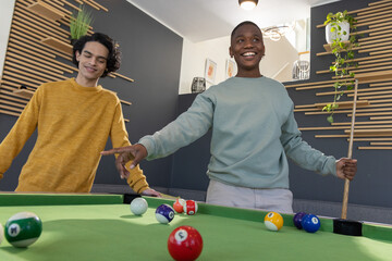 Obraz premium Diverse teenage male friends playing on green pool table in modern rec room with cue stick