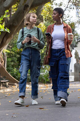 Diverse female friends walking on park path holding travel mug, coffee cup, smartphone, backpacks © wavebreak3