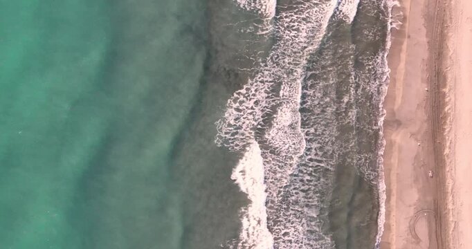 Aerial top down view of ocean waves rolling onto sandy beach with white foam and turquoise water