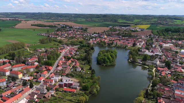 High-altitude aerial view of a small Czech town with red-roofed houses, a wooded island in a large pond, and patchwork green and brown farmland stretching to distant hills, Telc, Czech Republic