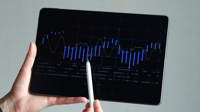 A person holds a tablet that displays a line and bar chart. The individual uses a stylus to interact with the data. Sunlight enters the space, highlighting the screen.