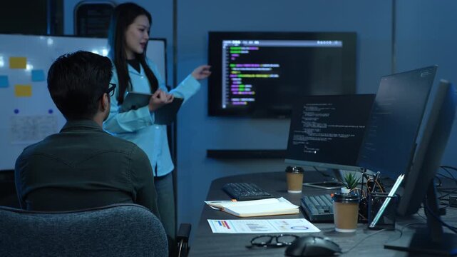 Asian female manager talking to male programmer writing code in dark blue office, Tech team discussing software development, cybersecurity and AI technology, Late night working business team concepts