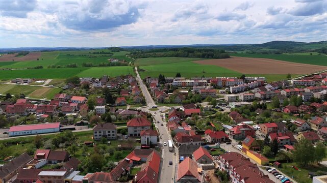 Aerial drone view of a small Czech town with a central roundabout, straight main road, red-roofed houses, and open green and brown farmland extending to wooded hills, Telc, Czech Republic