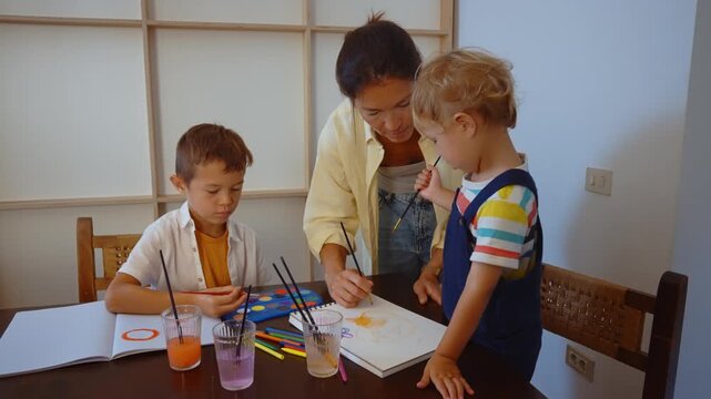Young creative mother and her two little sons having fun painting with watercolors at home, engaging in a family artistic activity that promotes bonding and creativity for the children