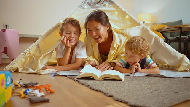 Cheerful mother reading a fairytale book to her two sons while lying inside a cozy teepee at home. Loving family enjoying a fun and educational activity together during a playful evening