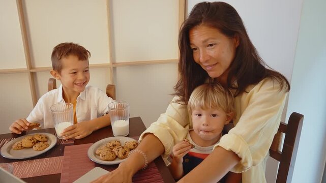 Happy family time with a mother and her two young sons sitting at the dining table, laughing and enjoying a snack of chocolate chip cookies and fresh milk, creating joyful childhood memories
