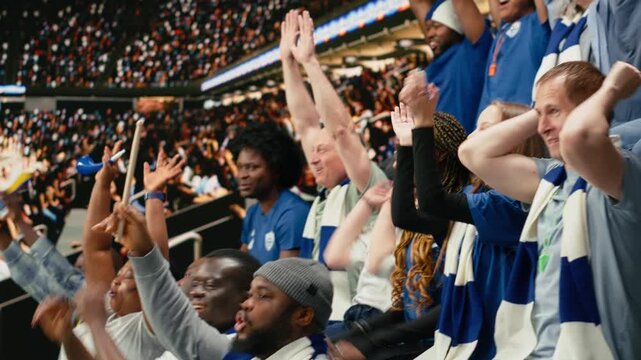 Blue team football supporters clapping and raising arms in crowded stands during exciting live soccer match with fan scarves and stadium atmosphere.