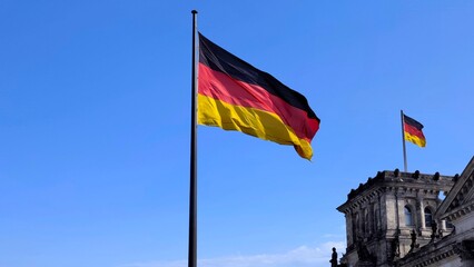 Photo of the German national flag waving proudly against a clear blue sky outside the historic Reichstag building in Berlin, Germany. © Antonios