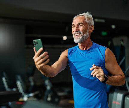 Portrait of a mature mid aged man using a phone and making a selfie photo, taking a break exercising in a gym, running using  thereadmill machine equipment, healthy lifestyle and cardio exercise at fi