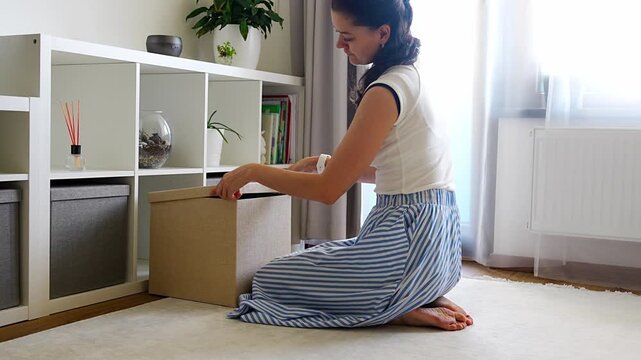 Young woman closing and putting away storage box after organizing belongings in modern home interior. Home organization and decluttering routine concept