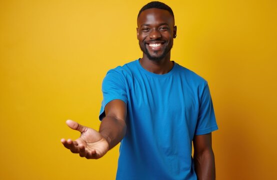 Young Black man smiles warmly extending open hand invitingly. He wears blue tee shirt against yellow backdrop. Gesture implies welcome helpfulness or offering something.