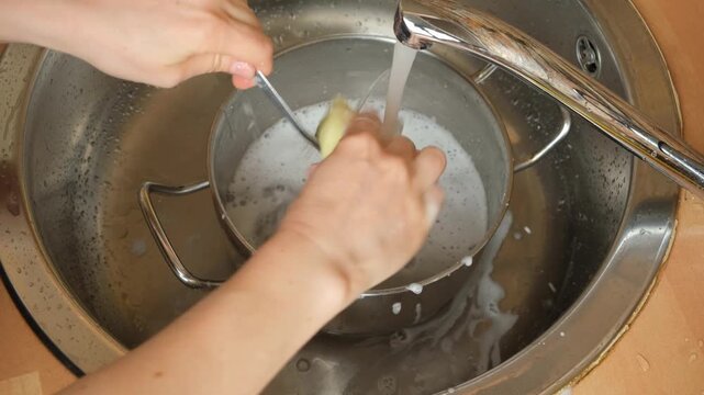 Hands washing and rinsing metal spoon with sponge over dirty stainless steel pot