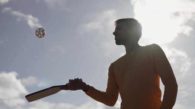 Slim man bouncing ball with wooden pickleball paddle in backlit light. Medium shot of athlete warming up before amateur padel tennis game in park against sunny sky.