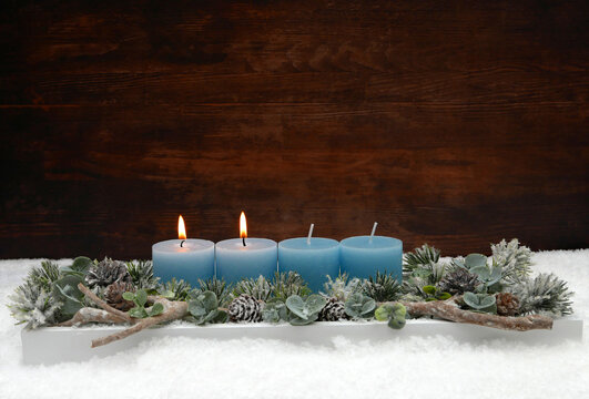 Zweiter Advent:  Adventsgesteck zum zweiten Advent   mit zwei brennenden blauen Kerzen, Eukalyptus und Tannenzweigen im Schnee vor einer Holzwand mit Textfreiraum.