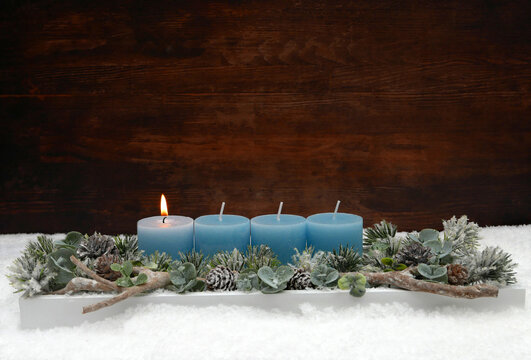 Erster Advent:  Adventsgesteck zum ersten Advent mit einer brennenden blauen Kerze, Eukalyptus und Tannenzweigen im Schnee vor einer Holzwand mit Textfreiraum.
