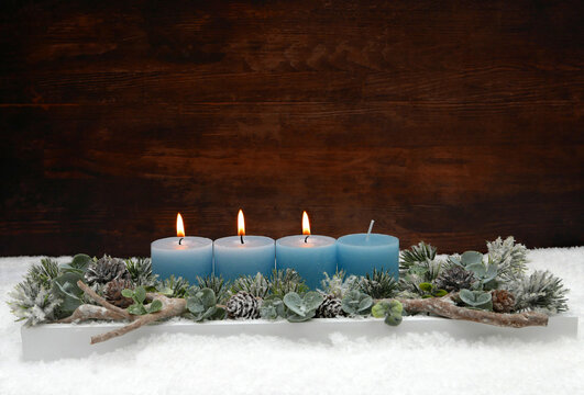Dritter Advent:  Adventsgesteck zum dritten Advent mit drei brennenden blauen Kerzen, Eukalyptus und Tannenzweigen im Schnee vor einer Holzwand mit Textfreiraum.