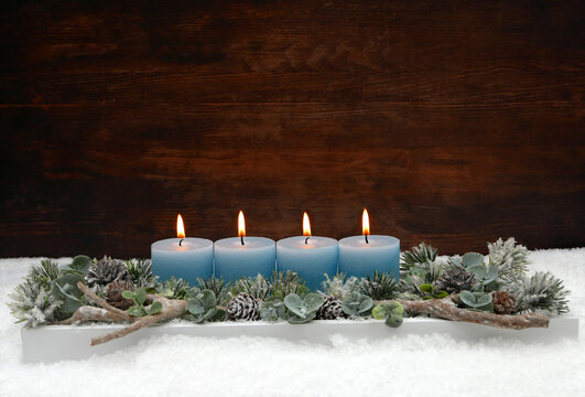 Vierter Advent:  Adventsgesteck zum vierten Advent mit vier brennenden blauen Kerzen, Eukalyptus und Tannenzweigen im Schnee vor einer Holzwand mit Textfreiraum.