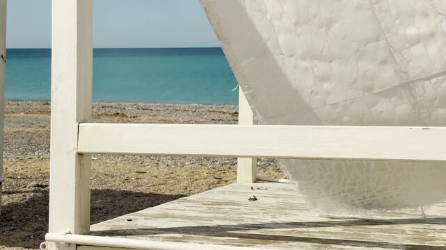A weathered wooden gazebo with frayed fabric sits peacefully on the sand. The calm blue sea and bright sunshine evoke a sense of quiet solitude and coastal relaxation.