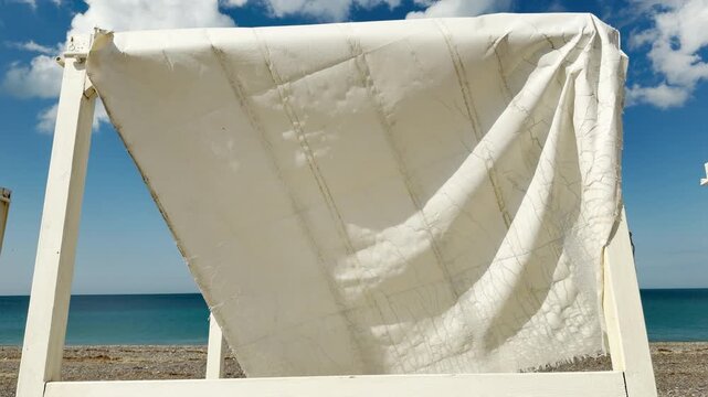 A cracked and frayed white canopy hangs heavily within a weathered wooden frame on a deserted beach. The quiet, empty seaside scene stirs a deeply nostalgic and peaceful feeling.