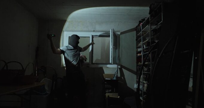Nighttime indoor scene of a burglar inside a basement room pointing toward an open window used for entry, illustrating home intrusion, break-in route, residential burglary