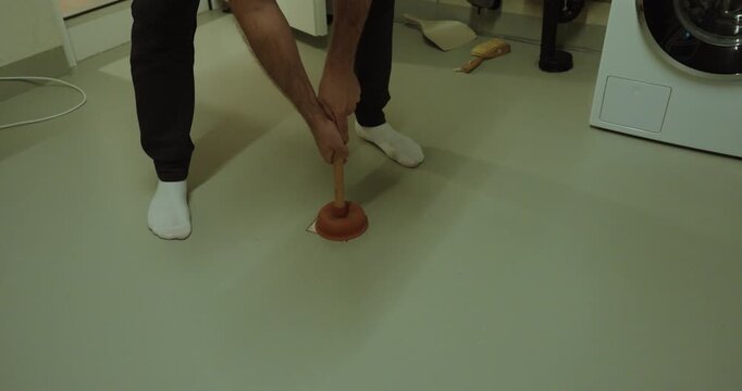 Indoor shot of a person using a plunger on a clogged floor drain, showing drainage problems, plumbing maintenance, blocked pipes, household repair, water flow issues, and home troubleshooting.