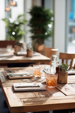 Wooden restaurant table with glass of iced tea and notebook setting. Cozy cafe interior with natural sunlight. Dining space atmosphere for lunch break or relaxed work environment.